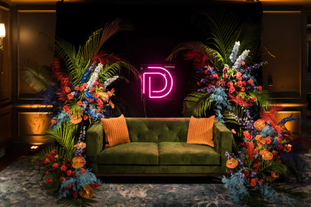 A plush green sofa flanked by vibrant floral arrangements with a neon letter 'd' on the wall behind it, situated in one of our expertly curated event venue spaces.